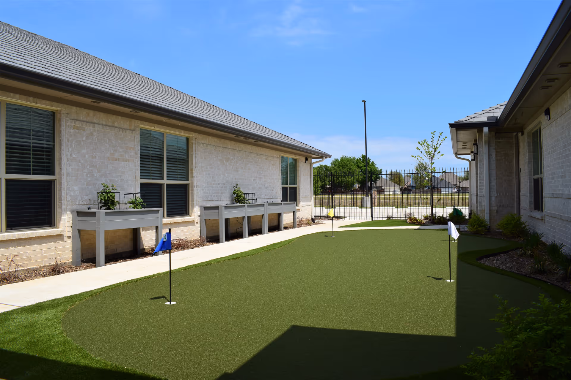 Outdoor courtyard area at Avalon Memory Care featuring a small putting green with three golf flags, surrounded by light-colored brick buildings with windows and raised planter boxes with plants. The area is enclosed by a black metal fence and has a clear blue sky overhead.