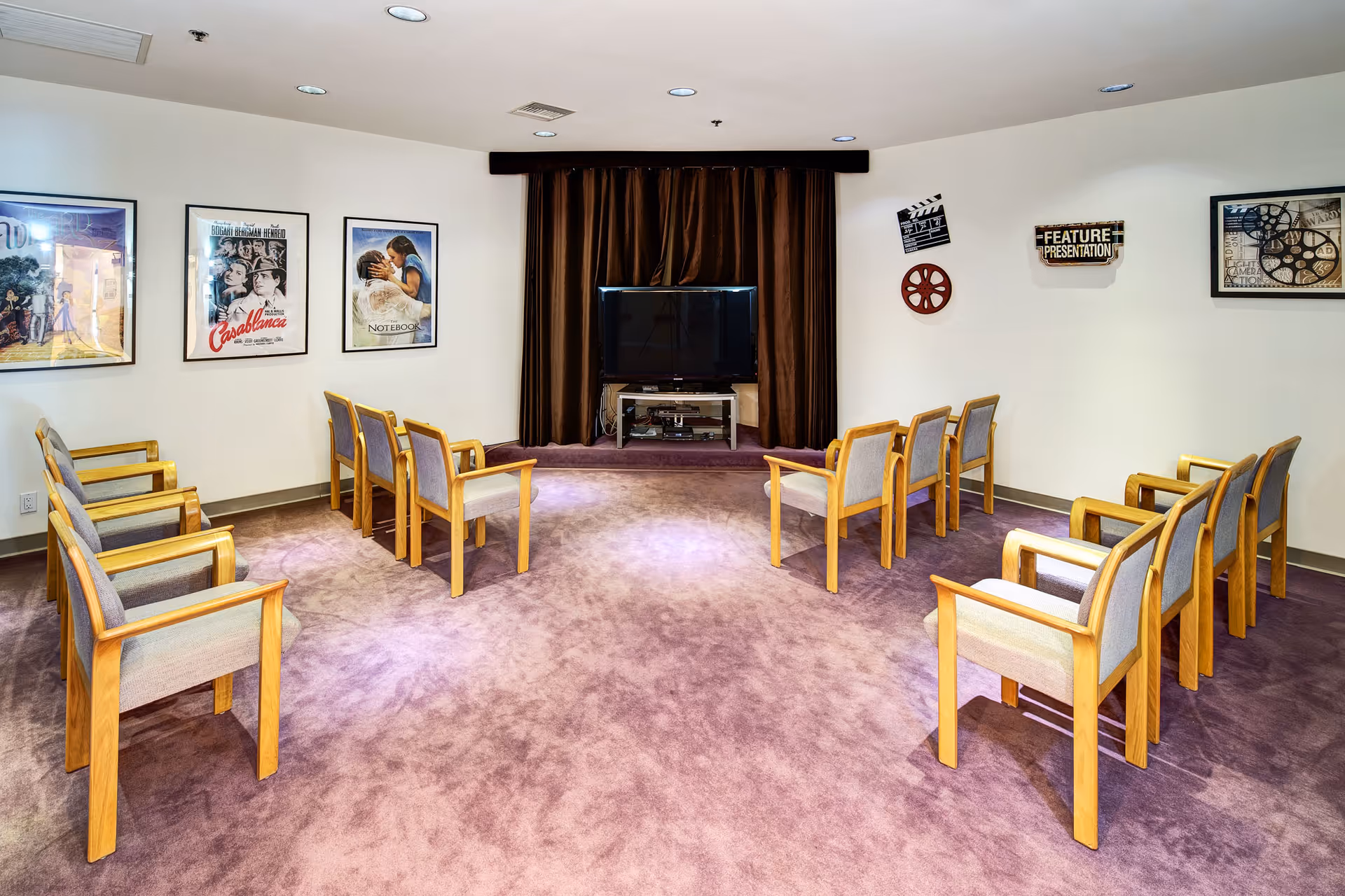A small movie screening room with two rows of wooden chairs with cushioned seats facing a flat-screen TV on a stand. The room has purple carpet and white walls decorated with framed classic movie posters and film-themed decorations.
