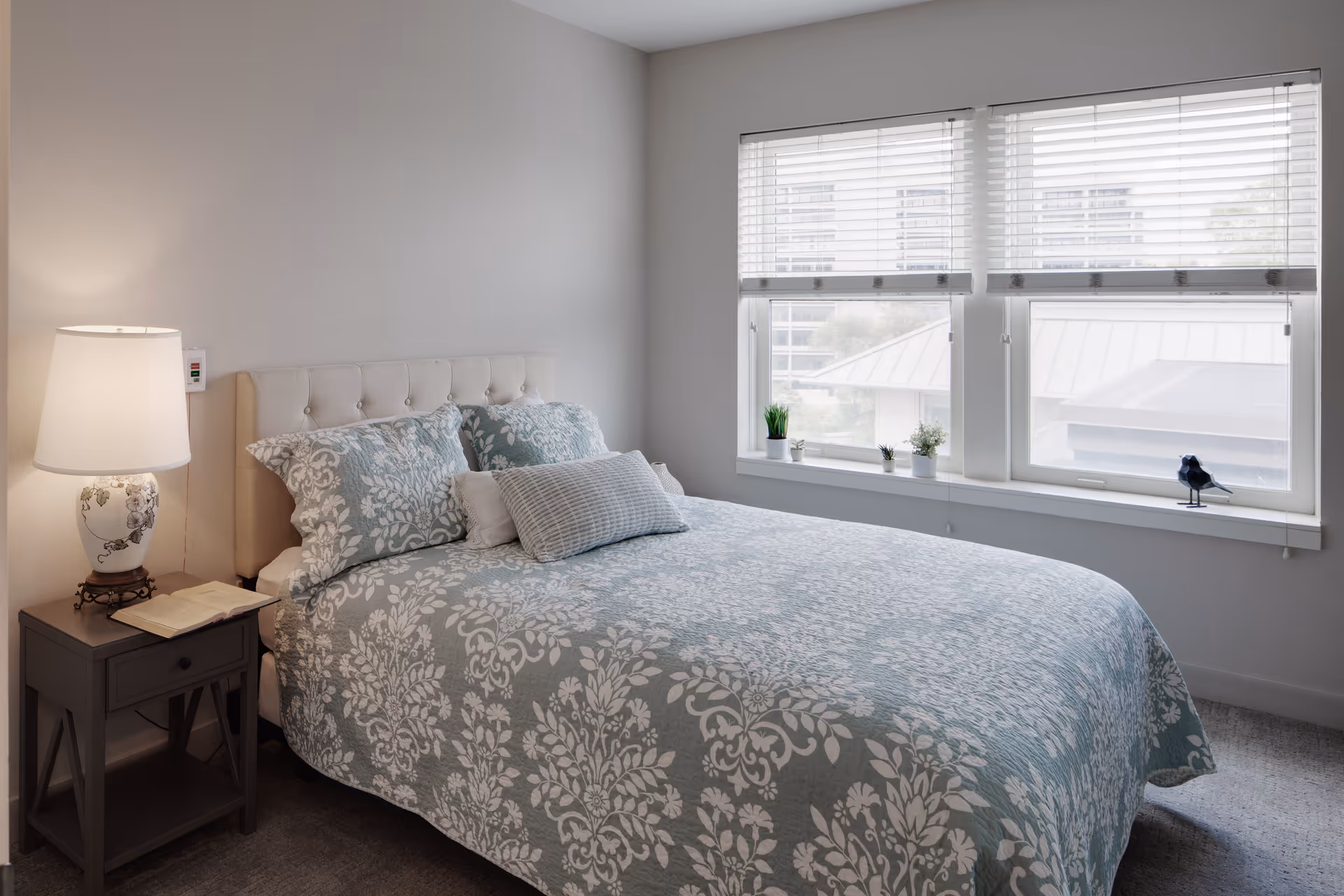 Bright bedroom with a patterned bed, tufted headboard, nightstand with a lamp and book, and three large windows with blinds.