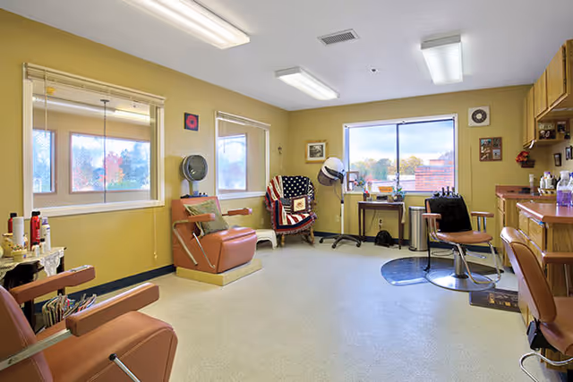 Interior view of a hair salon area in a senior living facility with multiple salon chairs, hair dryers, a window showing an outdoor view, and various hair care products on counters and shelves.