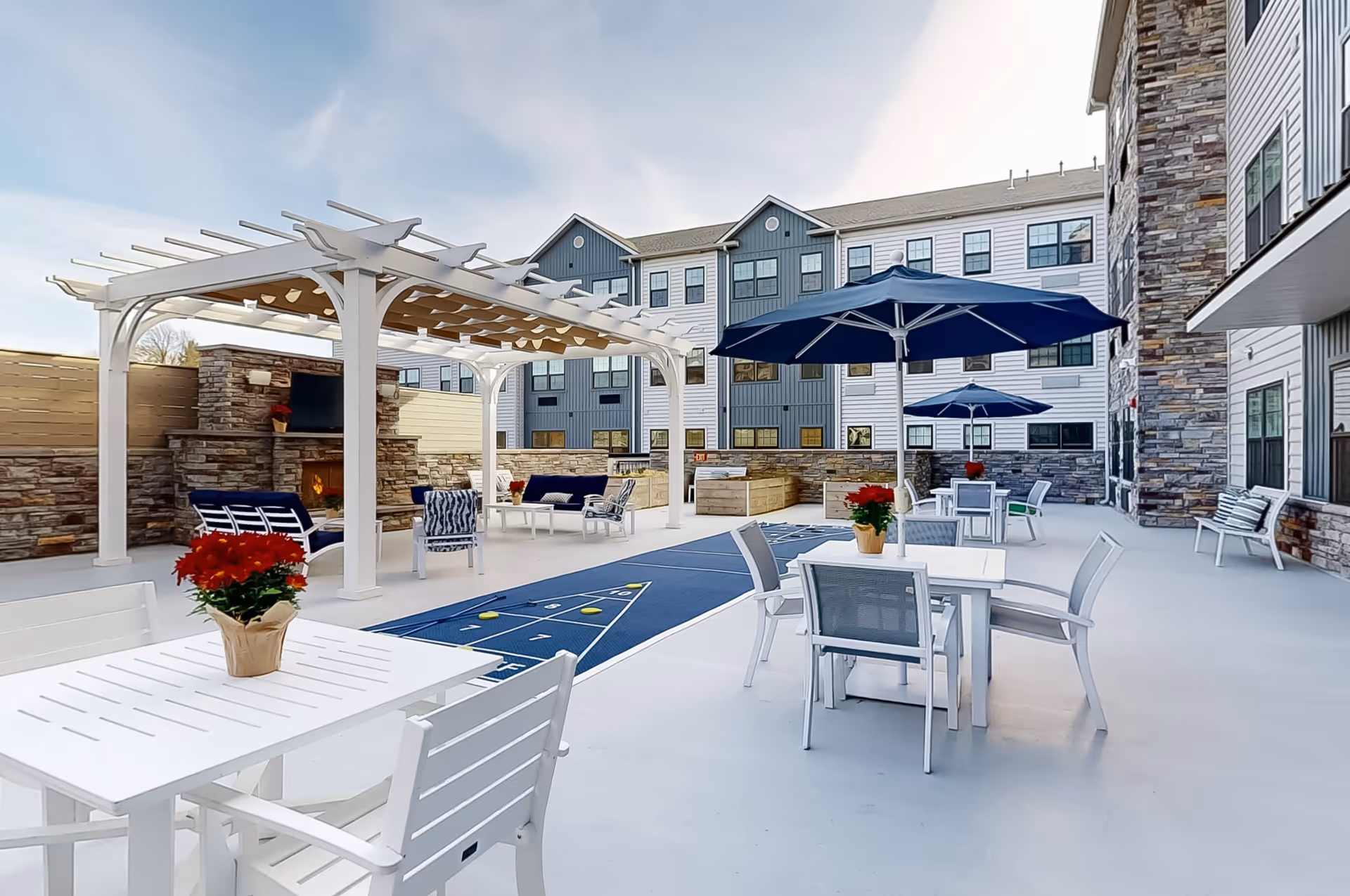 Outdoor patio area at Crescent Fields at Huntingdon Valley featuring white tables and chairs with potted red flowers, blue umbrellas, a shuffleboard court, a white pergola with seating underneath, and a stone fireplace mounted with a TV, all surrounded by a multi-story building with stone and siding exterior.