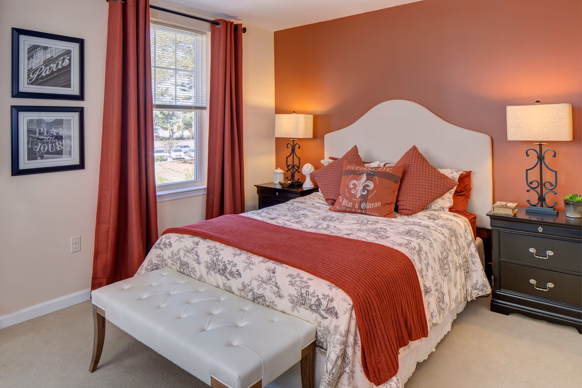 A cozy bedroom with a bed featuring a white and gray patterned bedspread and red throw blanket and pillows. The bed has a light-colored upholstered headboard. On either side of the bed are black nightstands with table lamps. A white tufted bench is placed at the foot of the bed. The walls are painted in a warm beige and reddish-brown color, with two framed pictures hanging next to a window with red curtains.