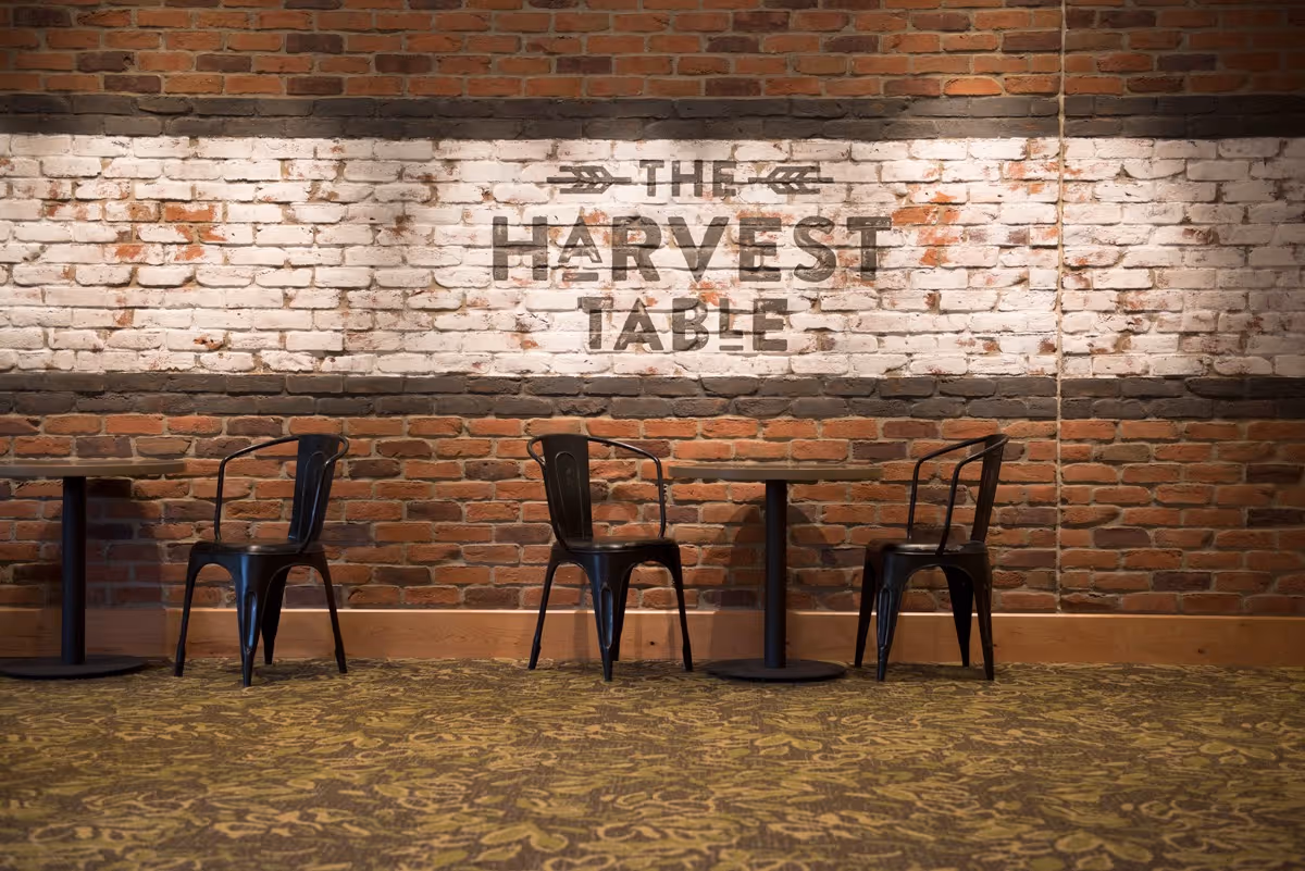 Interior view of a dining area with a brick wall featuring a white painted section with the words 'THE HARVEST TABLE'. There are three black metal chairs and two small round tables on a patterned carpeted floor.
