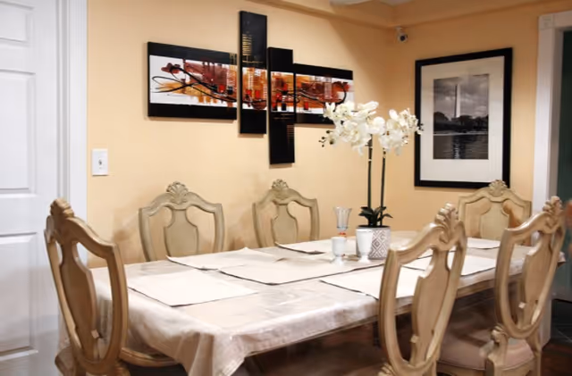 Dining room with a long table covered by a tablecloth, six ornate chairs, a vase of white flowers, and wall art.