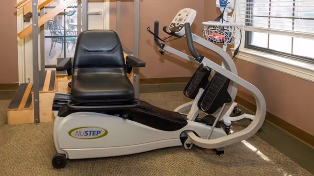 A NuStep exercise machine with a black seat and pedals in a room with carpeted floor, beige walls, a window with blinds, and a wooden handrail with stairs visible in the background.