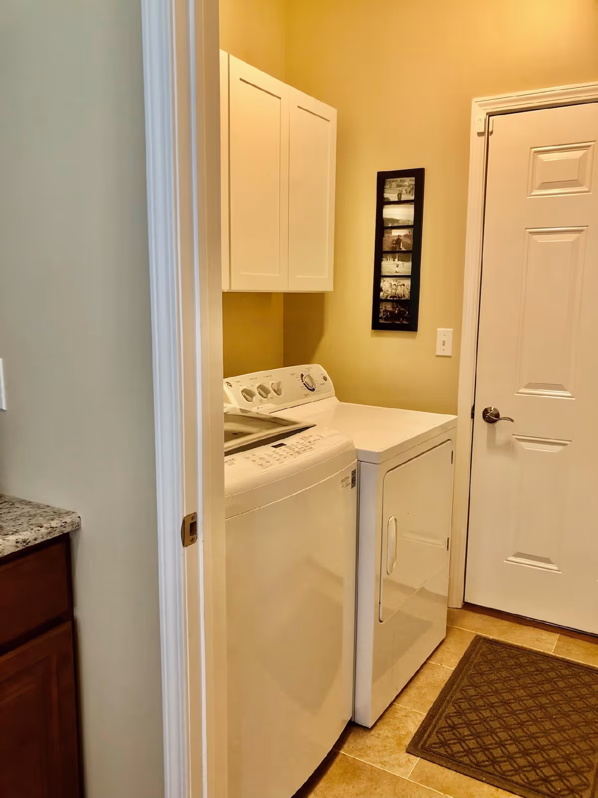 A laundry room with a white washing machine and dryer side by side, beige walls, a white cabinet mounted above the machines, a framed photo collage on the wall, a white door with a silver handle, and a brown floor mat on tiled flooring.