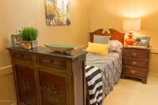 A cozy bedroom with a single bed covered in a gray and white striped blanket, a yellow pillow, and a black and white patterned pillow. Next to the bed is a wooden nightstand with a red lamp and a framed photo. In the foreground, there is a wooden dresser with decorative items including a green plant and a bowl. A painting hangs on the beige wall above the dresser.