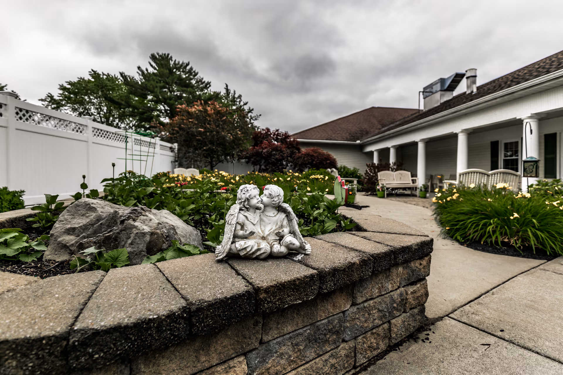 Outdoor garden area at Keepsake Village at Greenpoint featuring a stone planter with a small statue of two cherubs sitting together. The garden has various plants and flowers, with a white fence and trees in the background. A walkway leads to a building with a covered porch and benches under a cloudy sky.