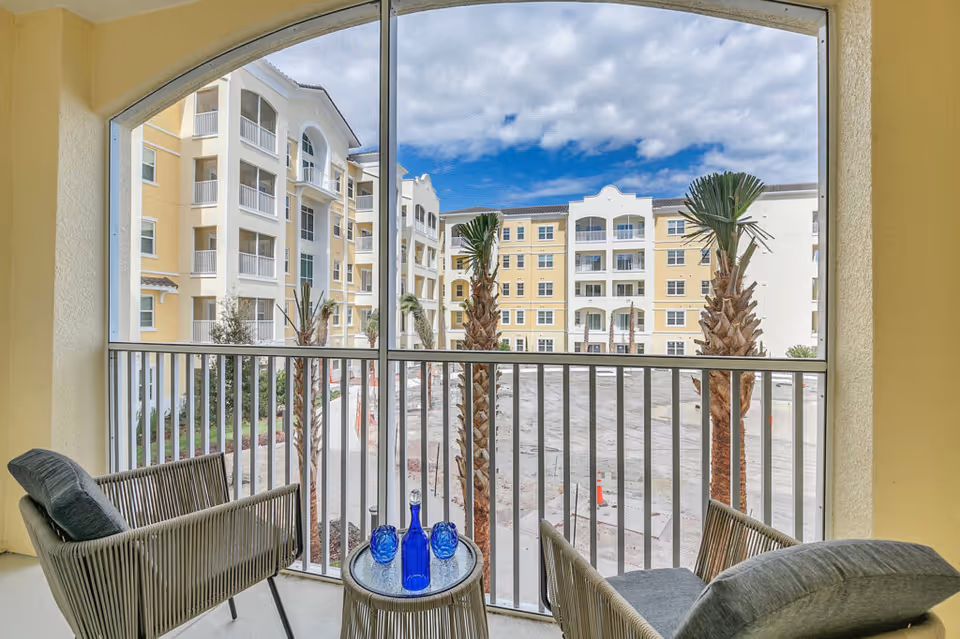 View from a screened balcony with two cushioned chairs and a small round table holding a blue bottle and two blue glasses, overlooking a courtyard with palm trees and a multi-story yellow and white building under a partly cloudy sky.