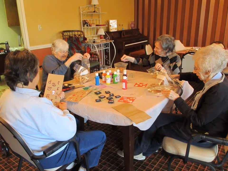 Four elderly women sitting around a table engaged in a craft activity with paint bottles, stamps, and paper bags in a cozy room with a piano and shelves in the background.