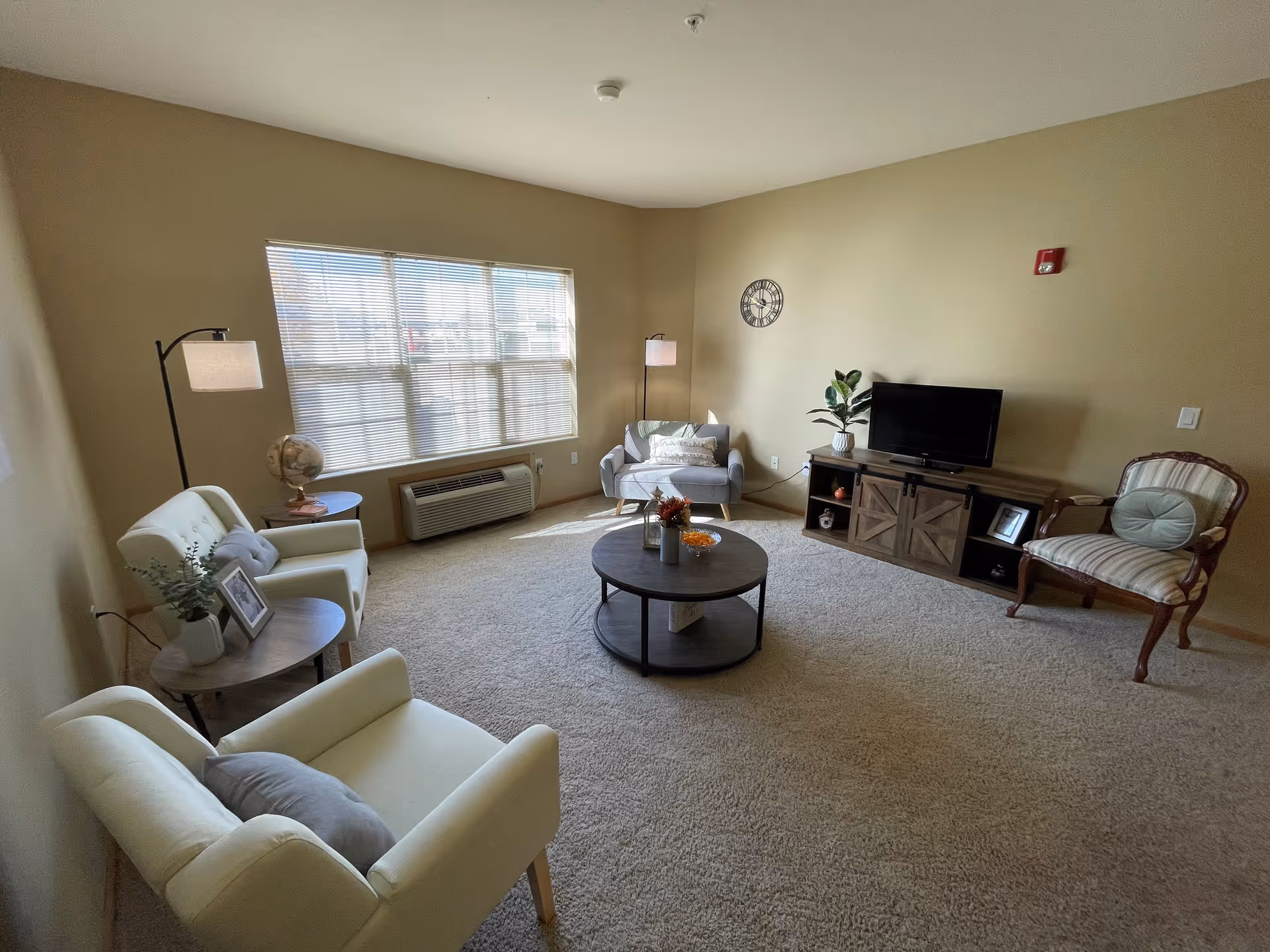 A cozy living room with beige walls and carpet. The room features a large window with blinds, two white armchairs with gray pillows, a small round wooden side table with a plant and framed photo, a floor lamp, a gray loveseat with pillows, a round wooden coffee table with decorative items, a wooden TV stand with a flat-screen TV, a potted plant, and a vintage-style armchair with striped upholstery and a round cushion. A wall clock is mounted on the wall above the loveseat.