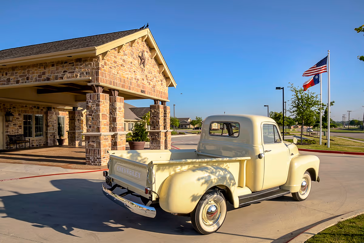 A vintage cream-colored Chevrolet pickup truck parked in front of a stone building with a covered entrance. The building has a star decoration on the gable and is surrounded by a paved driveway and landscaping. Two flags, including the American flag and the Texas state flag, are flying on flagpoles nearby under a clear blue sky.