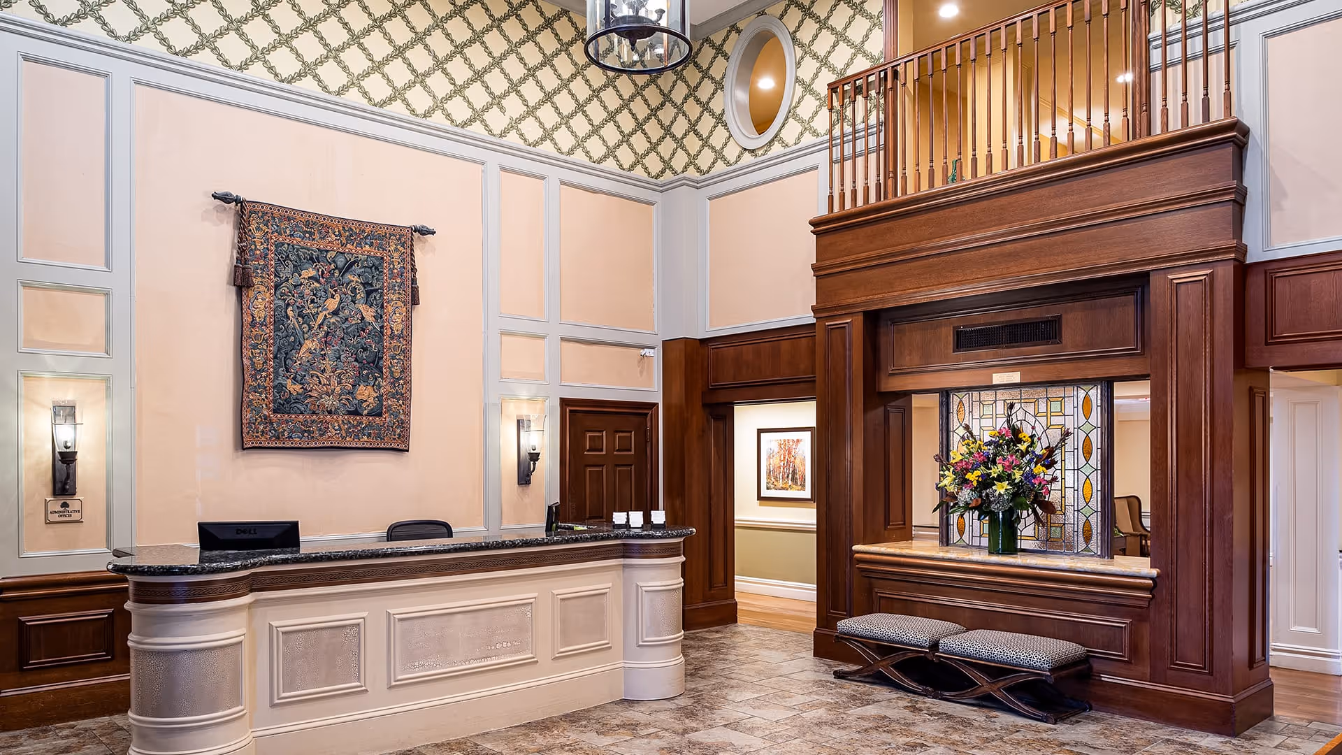 Reception area with a large front desk featuring a granite countertop, a decorative tapestry hanging on the wall behind it, and a wooden structure with a stained glass window and a vase of colorful flowers. The room has high ceilings with patterned wallpaper near the top and warm lighting fixtures on the walls.