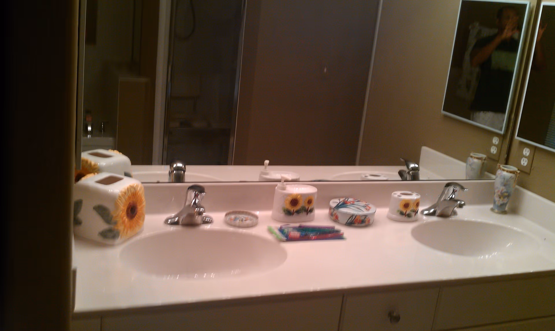 A bathroom countertop with two sinks and chrome faucets. On the counter are decorative containers with sunflower designs, a small round container with a floral pattern, a toothbrush holder with toothbrushes, and a floral vase. A large mirror spans the length of the countertop, reflecting the items and part of the bathroom. A person is partially visible in the mirror taking the photo.
