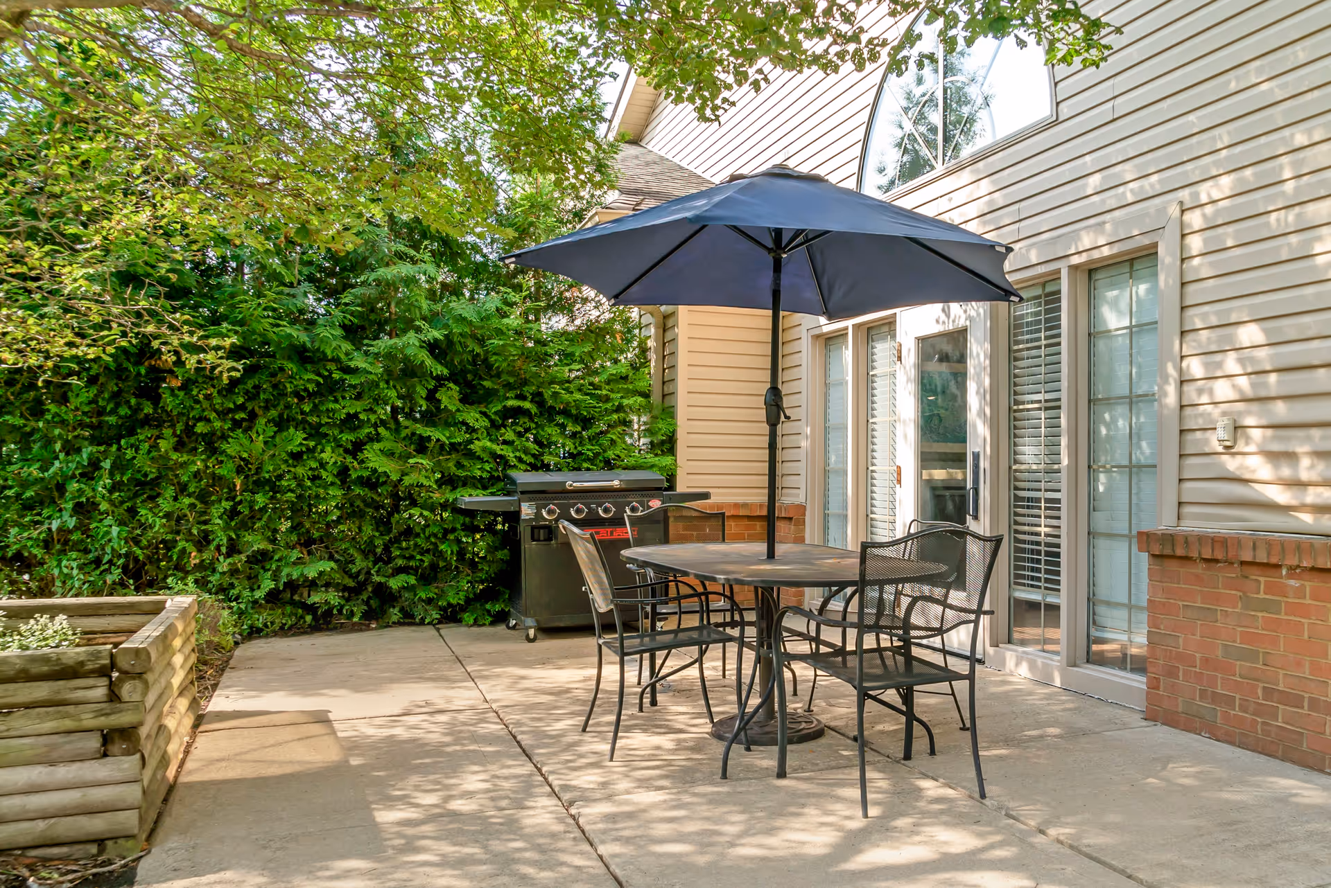 Outdoor patio area with a round metal table and four matching chairs under a large blue umbrella. There is a gas grill in the background near the house, which has beige siding and multiple glass doors and windows. The patio is surrounded by green bushes and trees providing shade.