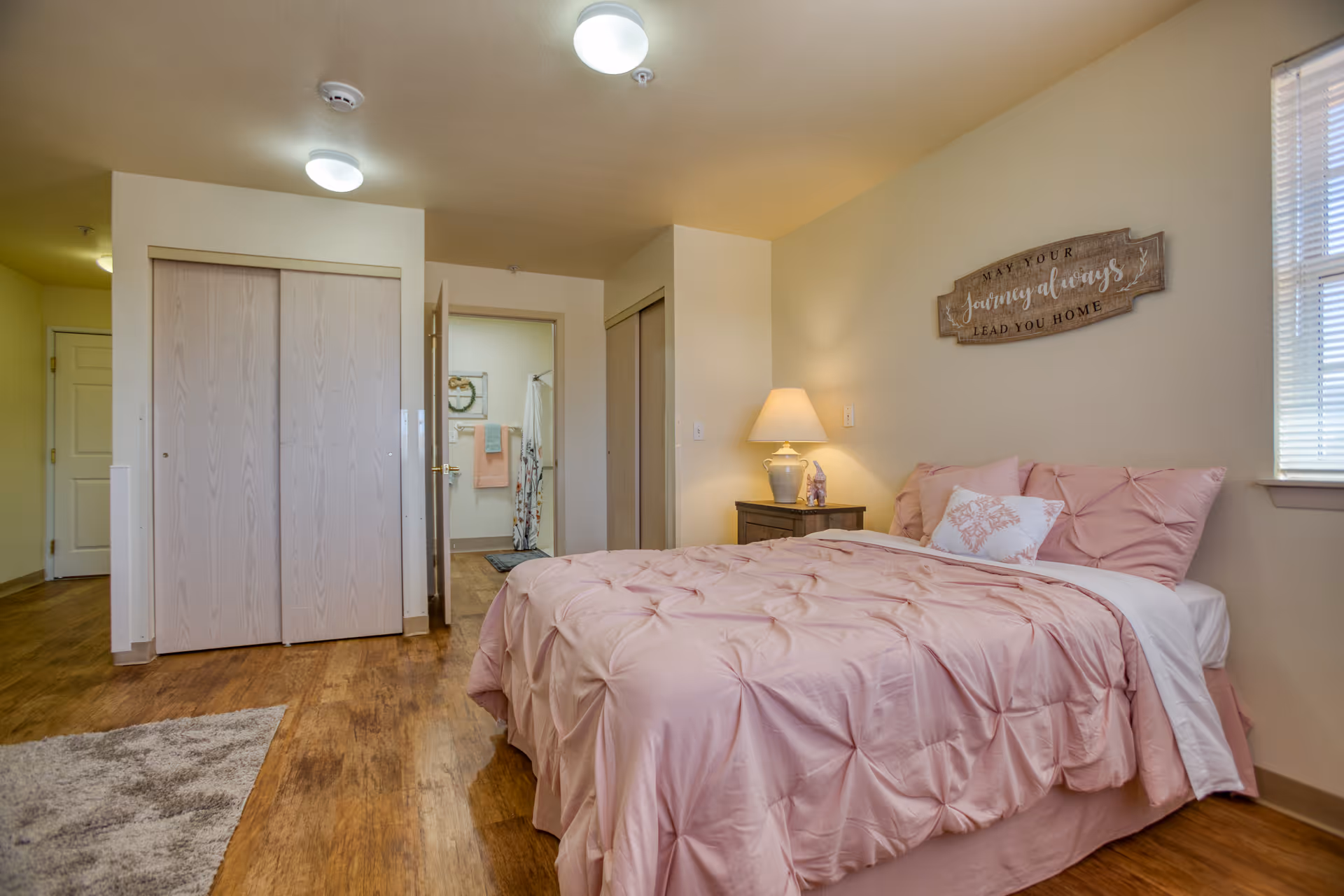 A cozy bedroom with a bed dressed in pink bedding and pillows. A wooden nightstand with a lamp and decorative items sits beside the bed. Above the bed is a wooden wall sign that reads 'May your journey always lead you home.' The room has wooden flooring, a window with blinds, and two closets with sliding doors. An open door reveals a glimpse of a bathroom with towels and a shower curtain.