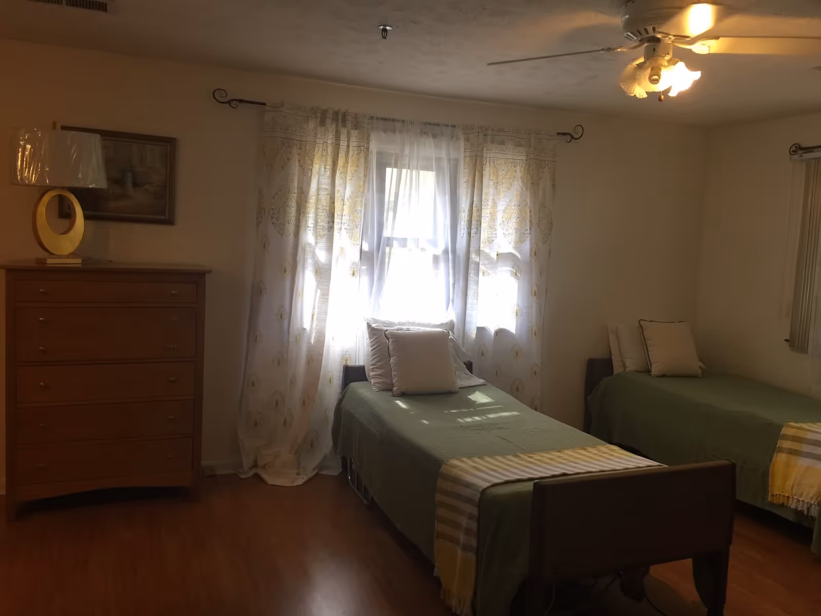 A bedroom with two single beds covered in green bedspreads and white pillows. There is a wooden dresser with a lamp and a framed picture on the wall above it. Sheer white curtains with a yellow pattern cover the window, and a ceiling fan with lights is visible on the ceiling.