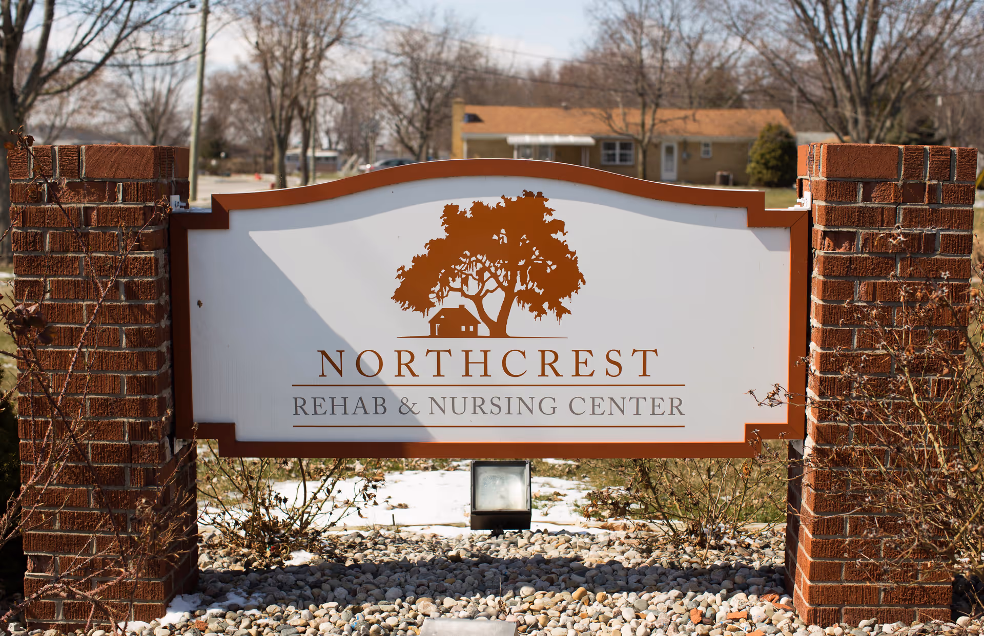 A sign for Northcrest Rehab & Nursing Center mounted between two brick pillars with a tree and house logo above the facility name. The background shows a residential area with leafless trees and a house.