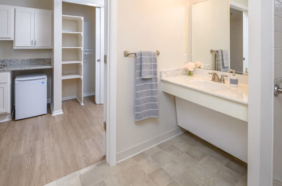 A clean bathroom with a large mirror above a white sink and countertop. A gray towel hangs on a towel rack on the wall. Adjacent to the bathroom is a small kitchen area with white cabinets, a granite countertop, and a mini refrigerator. There is an open doorway leading to a walk-in closet with shelves and hanging space. The bathroom floor is tiled, while the kitchen area has wood-style flooring.