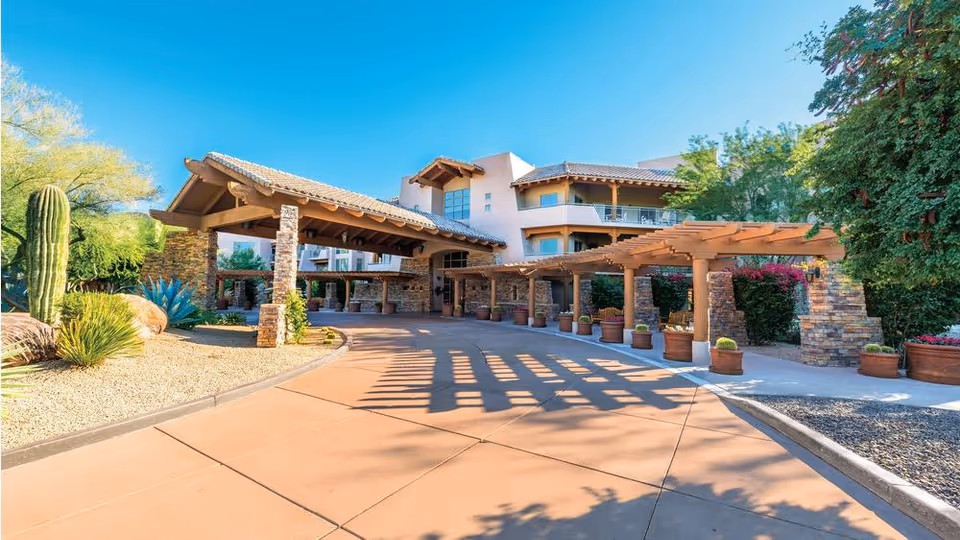 Exterior view of the entrance to Vi at Grayhawk senior living facility featuring a covered driveway with stone pillars, wooden pergola, desert landscaping with cacti and shrubs, and a clear blue sky.