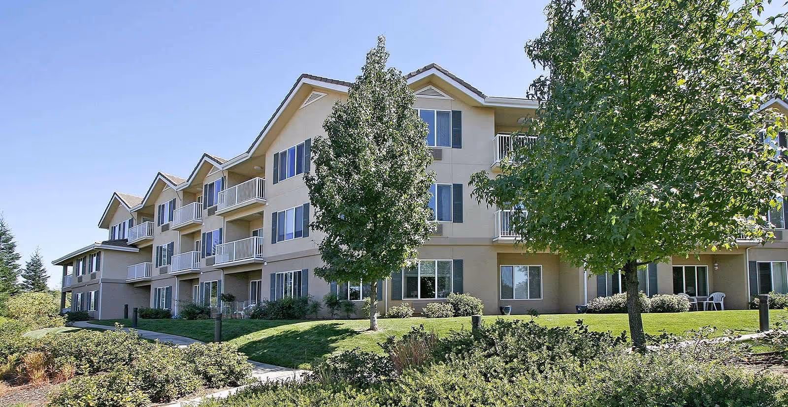 Exterior view of a three-story senior living facility building with beige walls and multiple balconies. The building is surrounded by green grass, trees, and shrubs under a clear blue sky.