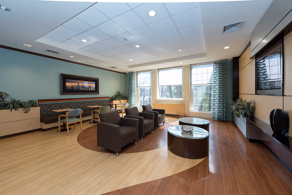 A bright and spacious common area with large windows letting in natural light. The room features a seating area with four dark armchairs arranged around two round glass-top coffee tables. There is a built-in bench with patterned upholstery along one wall accompanied by small tables and chairs. The walls are decorated with plants and a framed panoramic cityscape photograph. The floor has a two-tone wood pattern, and a large flat-screen TV is mounted on a wood-paneled wall.