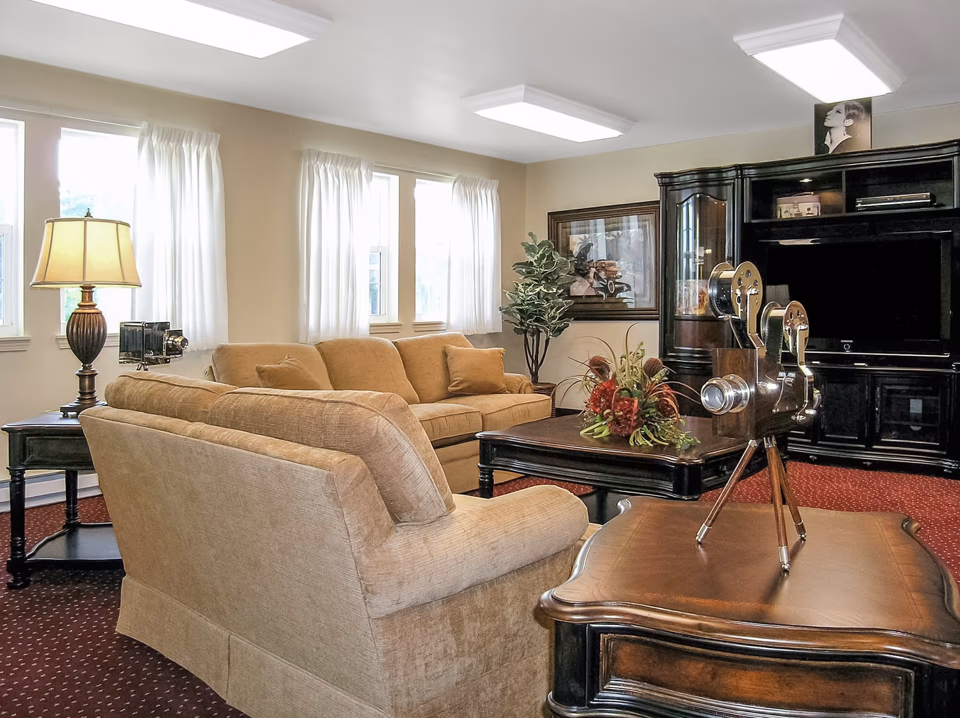 A cozy living room with beige sofas, a wooden coffee table with a floral arrangement, a side table with a lamp, and a large dark wood entertainment center with a TV. The room has multiple windows with white curtains and a red carpeted floor.