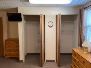 Interior view of a room with two open closets side by side, a wall clock mounted between them, a wooden dresser with a lamp on the right side near a window with curtains, and another wooden dresser with a television mounted above it on the left side.