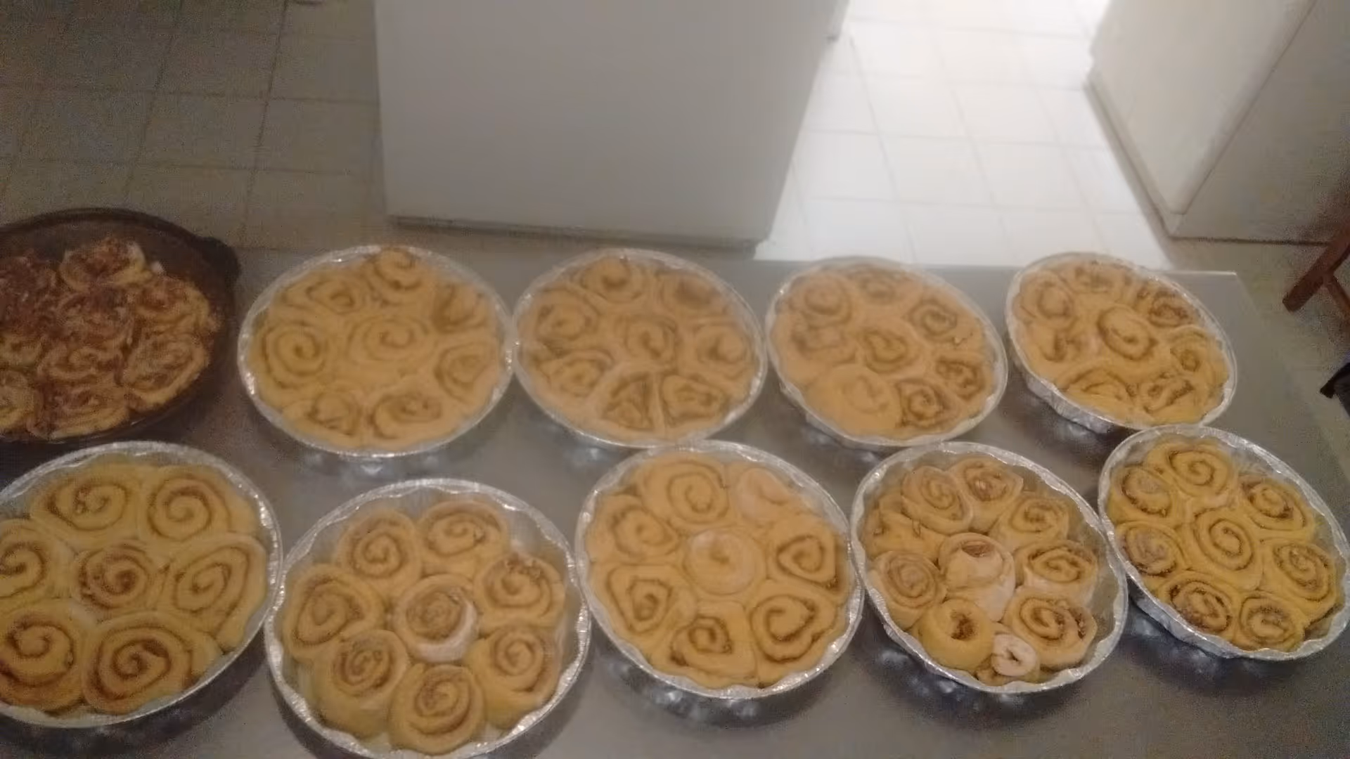 Several aluminum pans filled with unbaked cinnamon rolls arranged on a kitchen counter in a tiled kitchen.