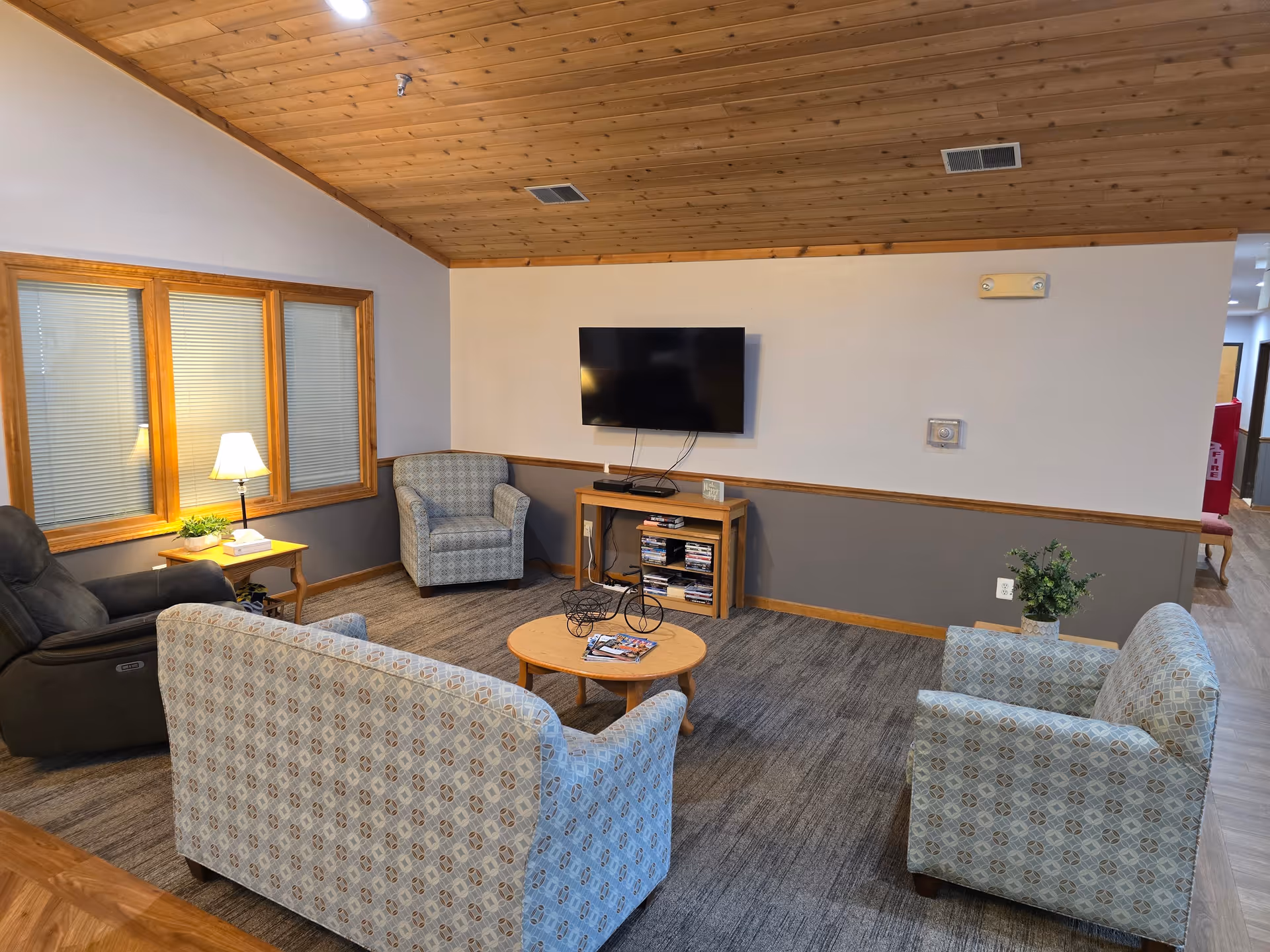 A cozy living room area with a wooden ceiling, carpeted floor, and neutral-colored walls. The room features a wall-mounted flat-screen TV above a wooden media console with DVDs, a round wooden coffee table with magazines, and four upholstered chairs arranged around the table. A side table with a lamp and a small plant is placed near a window with blinds.
