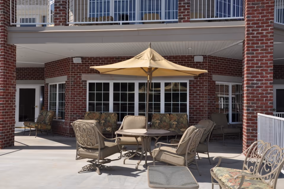 Outdoor patio area with cushioned metal chairs and a round table under a large beige umbrella, surrounded by a brick building with large windows and a balcony above.