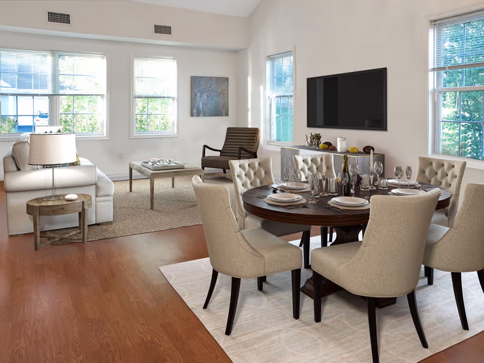 A bright and spacious living and dining area with large windows letting in natural light. The dining area features a round wooden table set with plates, glasses, and silverware, surrounded by six upholstered chairs. The living area includes a beige sofa, a wooden side table with a lamp, a coffee table, and a modern armchair. A flat-screen TV is mounted on the wall above a console table with decorative items.