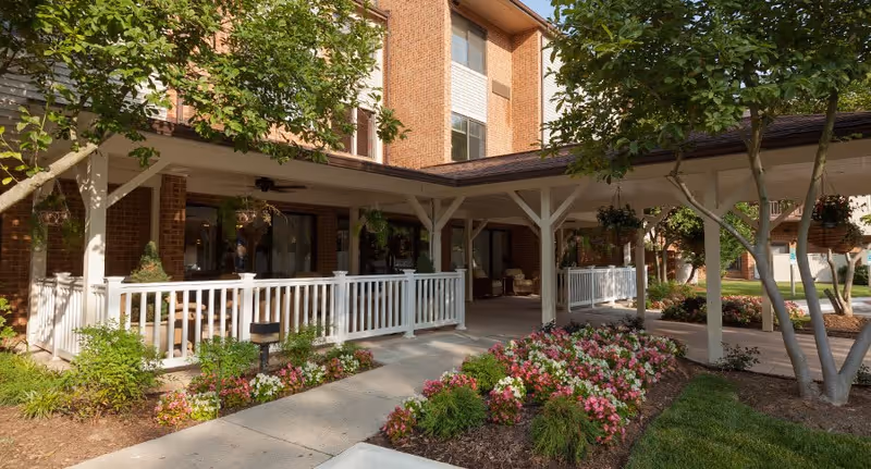 Outdoor view of Potomac Place Assisted Living and Memory Care showing a brick building with a covered walkway supported by white wooden beams. There are flower beds with pink and white flowers along the walkway, green shrubs, and trees providing shade.