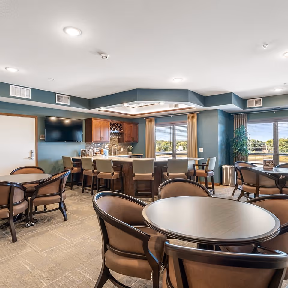 Well-lit communal dining and lounge area with round tables and chairs and a central bar counter with stools by large windows.