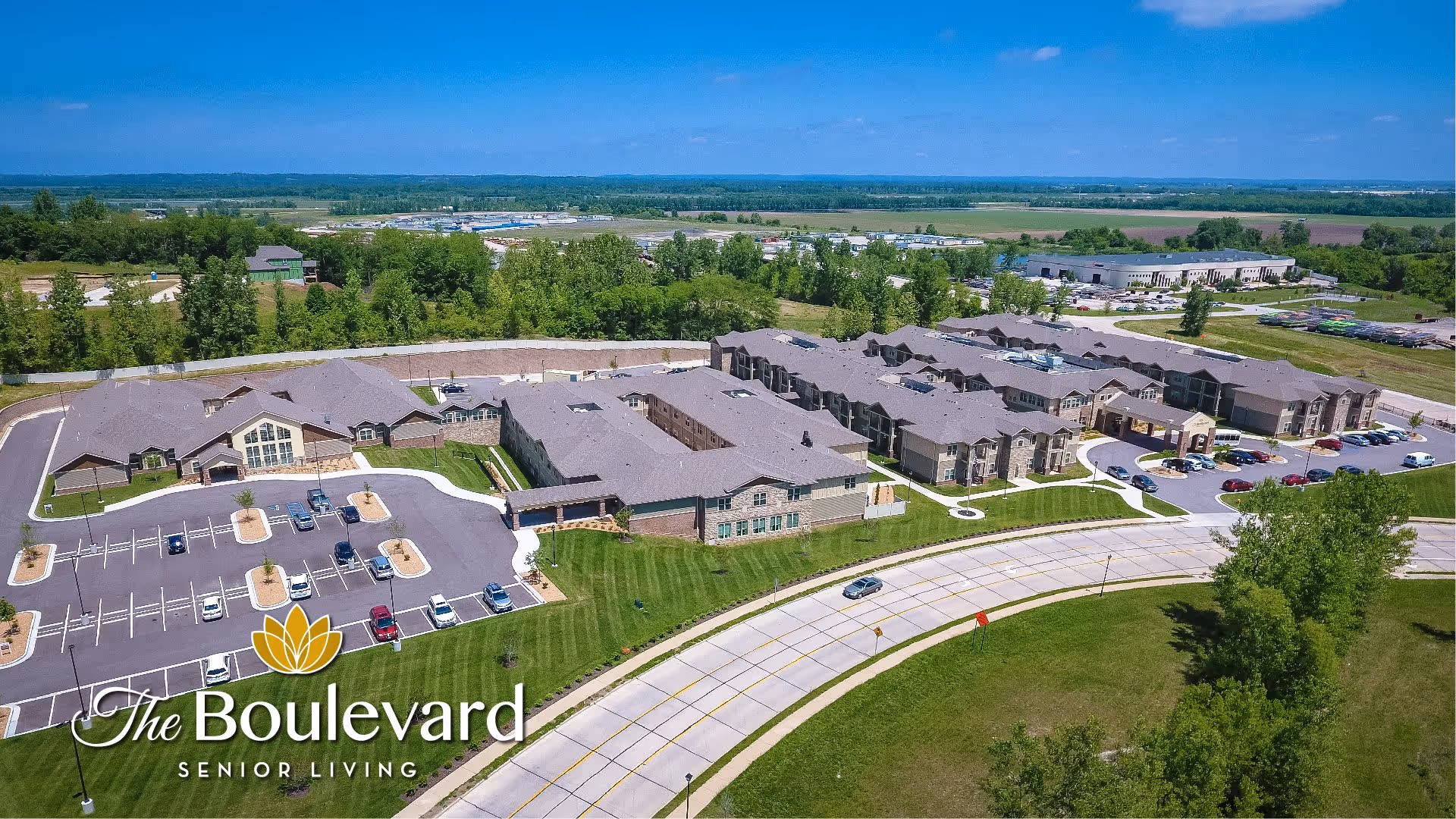 Aerial view of The Boulevard of St. Charles Senior Living facility showing multiple connected buildings with gray roofs, parking lots with cars, surrounding green lawns, trees, and a curved road in front. The landscape extends to a distant horizon under a clear blue sky.