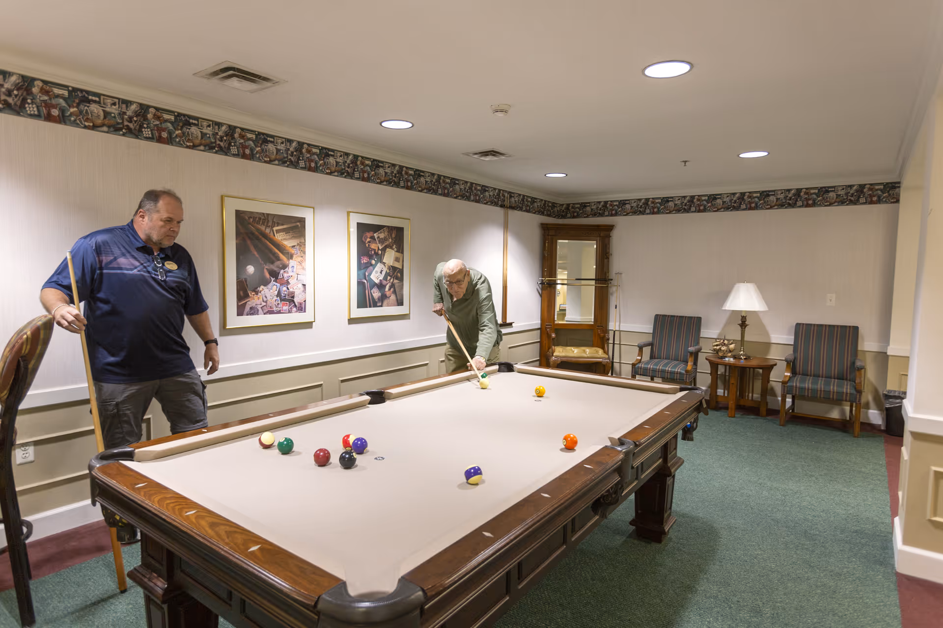 Two men play billiards at a pool table in a carpeted recreational room with chairs, framed artwork, and a lamp.