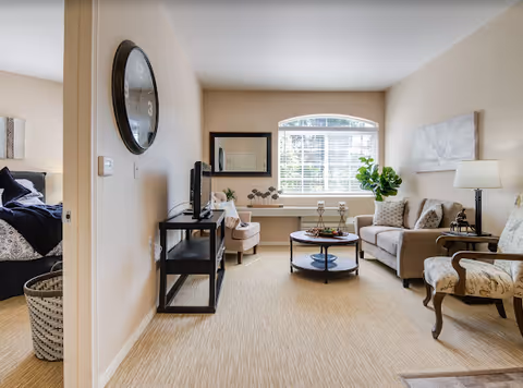 A bright and cozy living room with beige walls and carpet. The room features a beige sofa with patterned pillows, a round coffee table with decorative items, an armchair with a floral pattern, a side table with a lamp, and a TV on a black metal stand. A large window with white blinds lets in natural light, and a large mirror hangs on the wall near the window. A glimpse of a bedroom with a bed and dark bedding is visible through an open doorway.