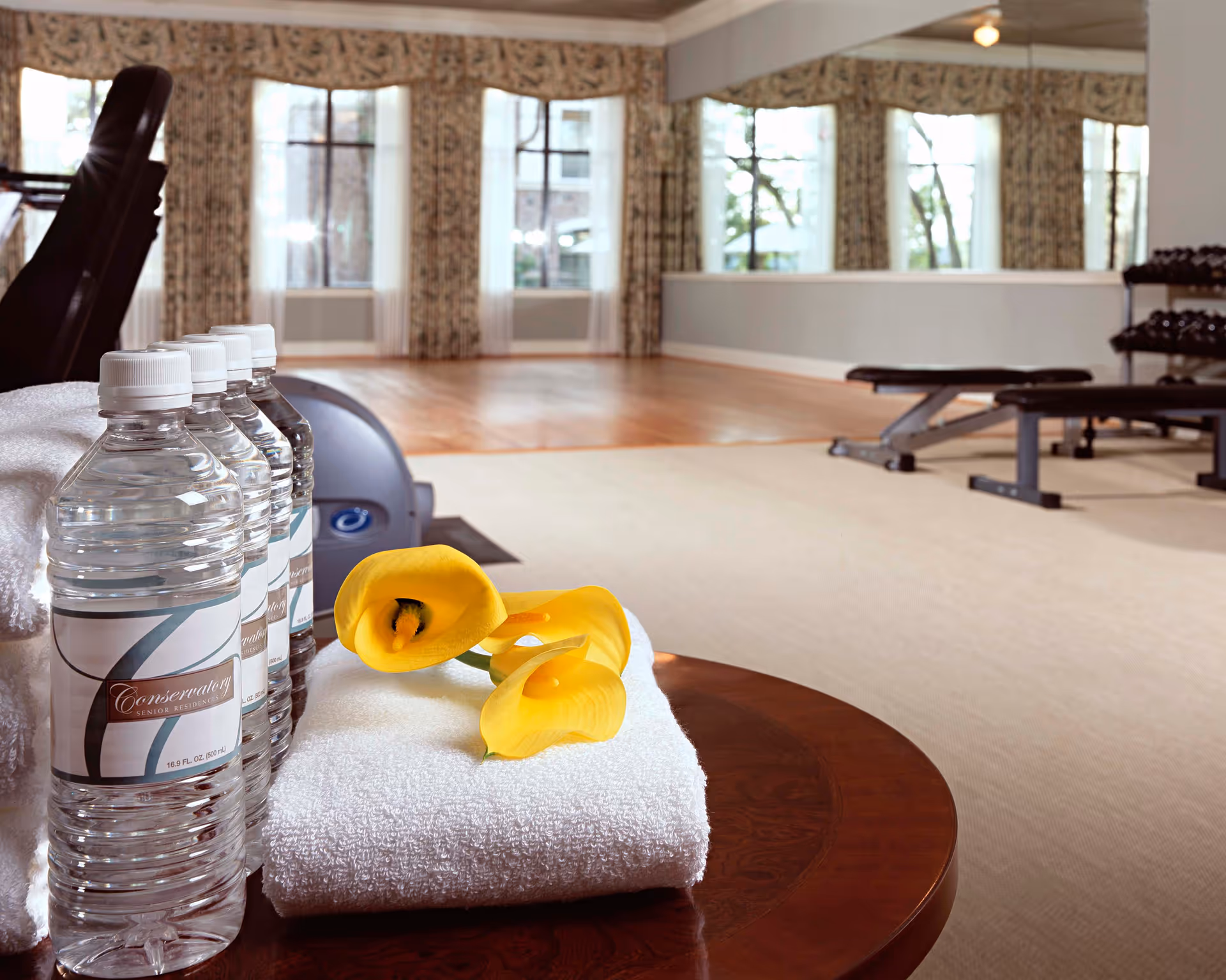 A fitness room with exercise equipment including a bench and dumbbells. In the foreground, there are several bottles of water labeled 'Conservatory Senior Residence' placed on a white towel with three yellow calla lilies on top. Large windows with patterned curtains and a mirrored wall are visible in the background.