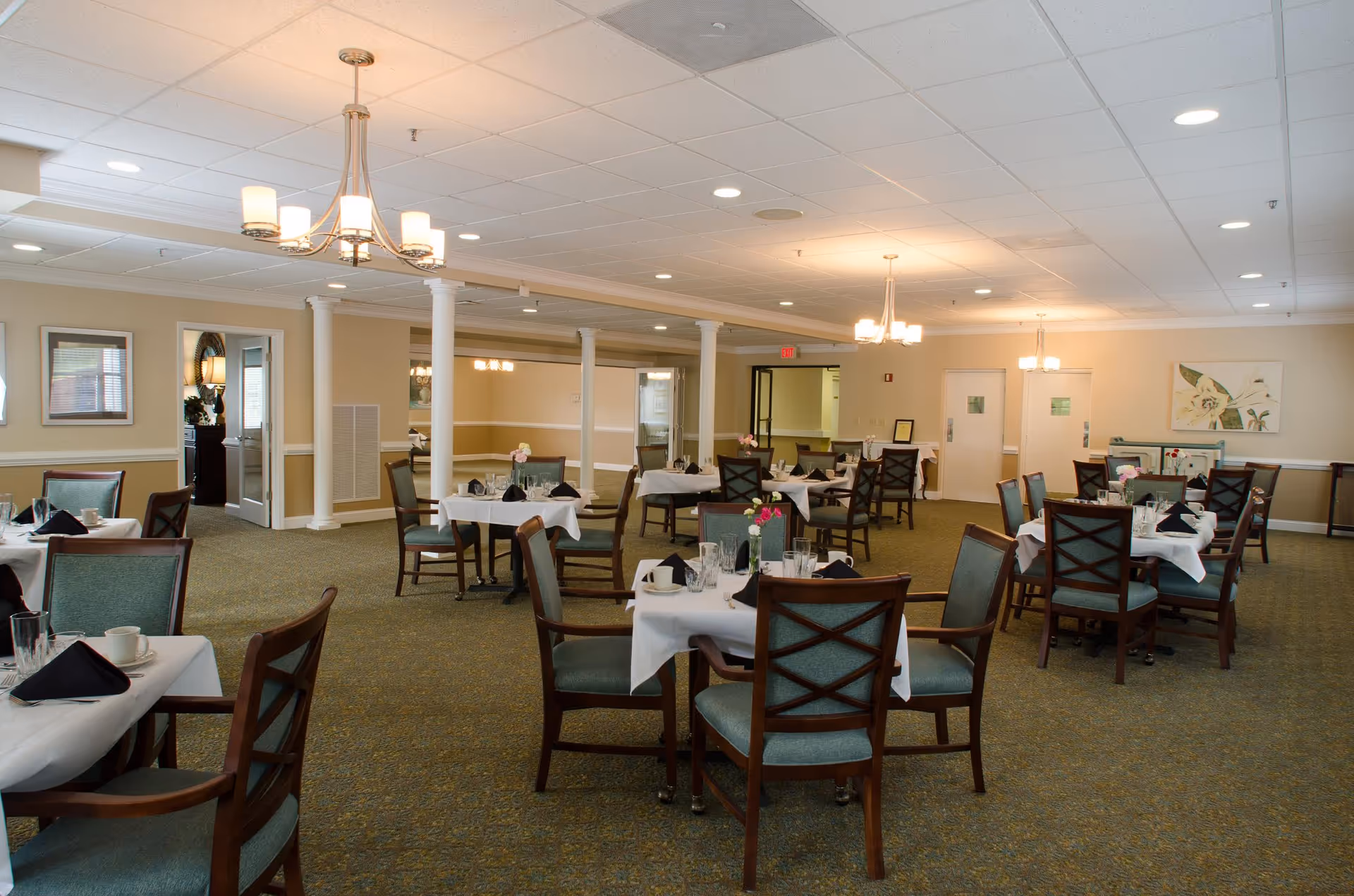Spacious senior living dining room with multiple round tables set with white tablecloths, chairs, and overhead chandeliers.