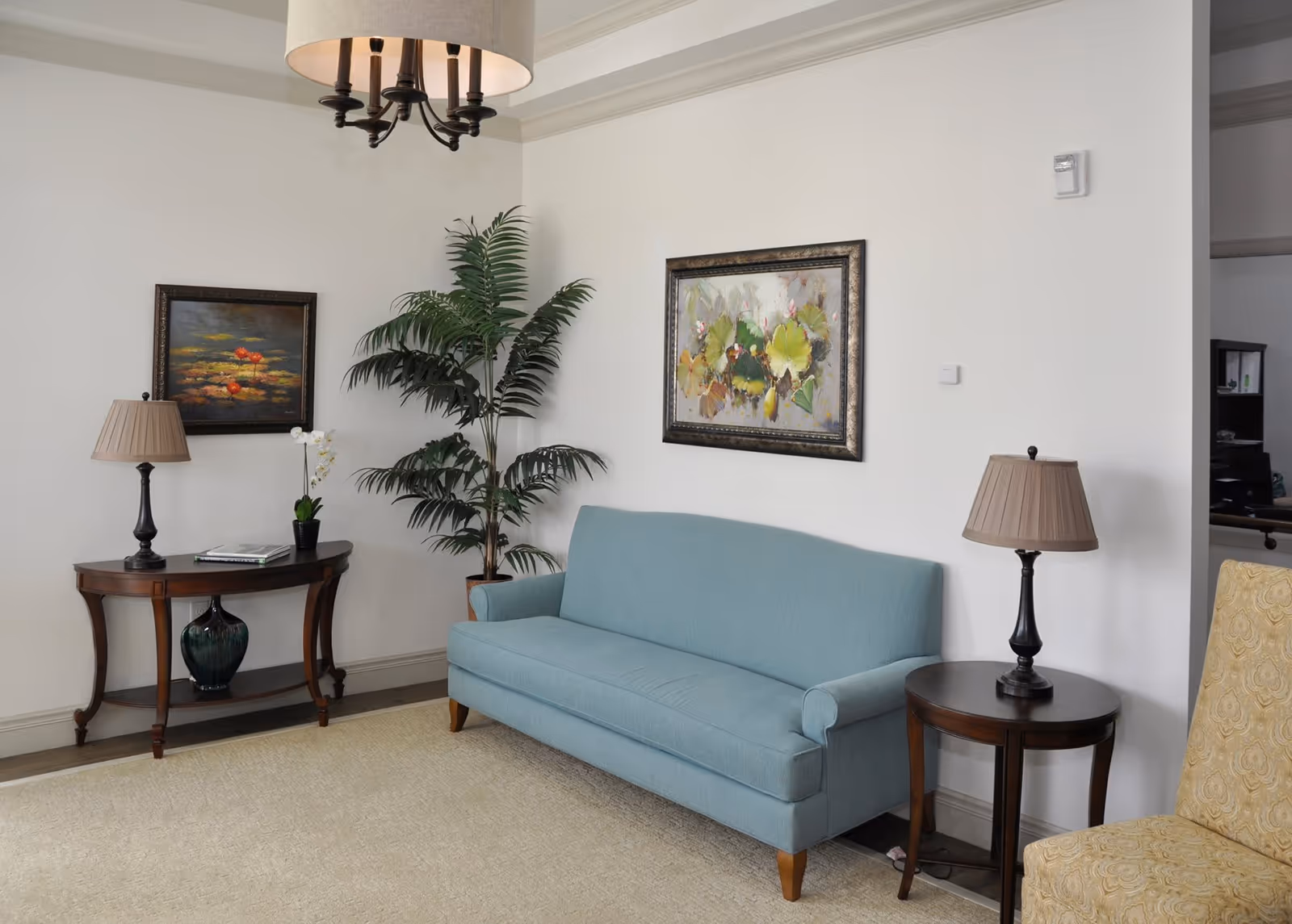 A cozy living room with a light blue sofa, a wooden side table with a lamp, a wooden console table with a lamp and a small plant, two framed paintings on the white walls, a large potted plant in the corner, and a beige patterned armchair partially visible.