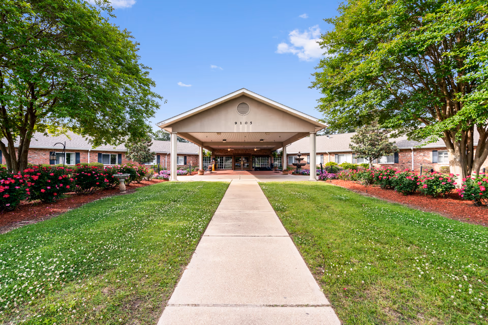Front exterior view of Southern Hills Healthcare and Rehabilitation building with a covered entrance, a concrete walkway leading up to it, green lawns, blooming pink flowers, and trees on both sides under a blue sky.