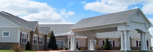 Exterior view of a senior living facility building with a covered entrance supported by white columns, beige siding, and a gray shingled roof under a partly cloudy sky.