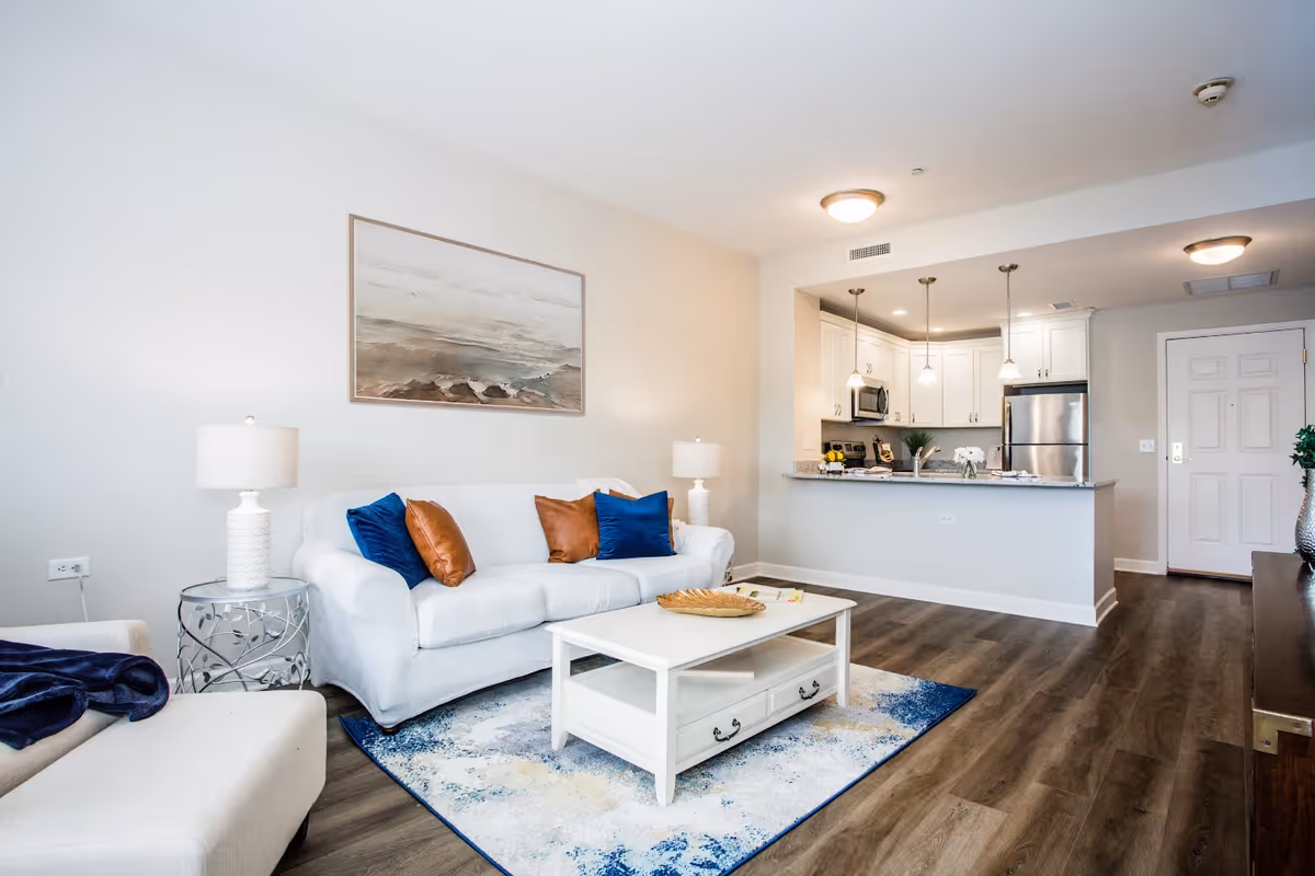 A bright and modern living room with a white sofa adorned with blue and brown pillows, a white coffee table on a blue and white patterned rug, a side table with a lamp, and an open kitchen area with white cabinets, stainless steel appliances, and pendant lights.