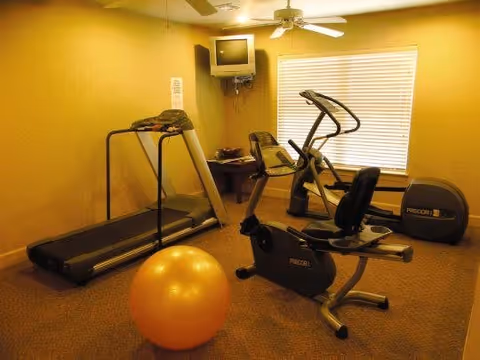 Small fitness room with a treadmill, stationary bike, elliptical machine, and a large orange exercise ball. A small TV is mounted in the corner above a table with some items on it. The room has beige walls and carpet, with a window covered by blinds.
