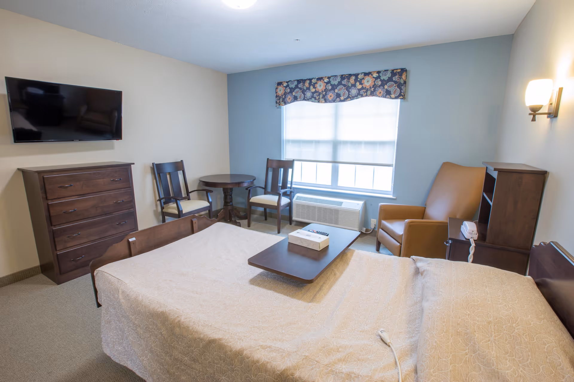 A well-lit bedroom in a senior living facility with a bed covered in a beige patterned bedspread, a wooden dresser with a flat-screen TV mounted above it, two wooden chairs and a round table near a window with a floral valance, a brown armchair, and a small wooden shelving unit with a telephone on it.