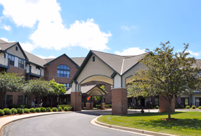 Exterior view of a senior living facility building with a covered entrance, brick pillars, and a curved driveway. The building has multiple windows, a sloped roof, and is surrounded by green landscaping and trees under a partly cloudy sky.
