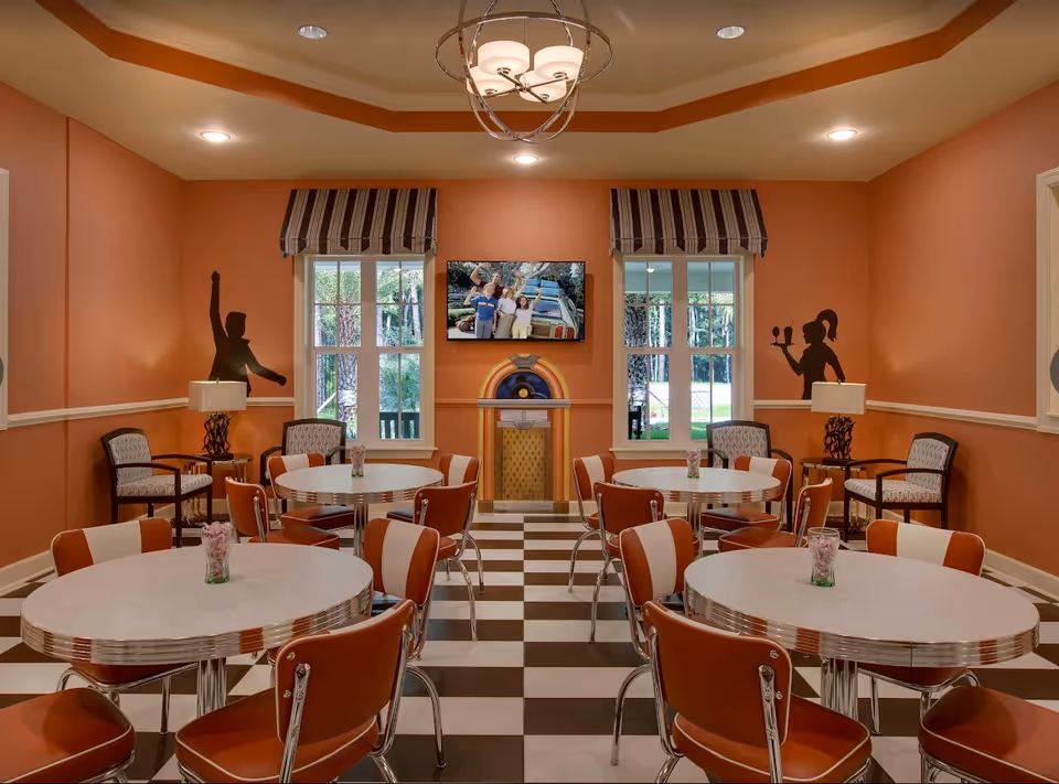 A retro-style dining room with orange walls and a checkered black and white floor. The room features round tables with orange and white chairs, a ceiling light fixture, two windows with striped valances, a wall-mounted TV showing a family photo, and silhouettes of people on the walls. There are also two armchairs with side tables and lamps in the corners.