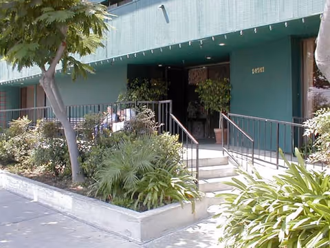 Entrance to a building with green exterior walls, a small staircase with handrails leading up to a door, and various plants and shrubs surrounding the entrance area.