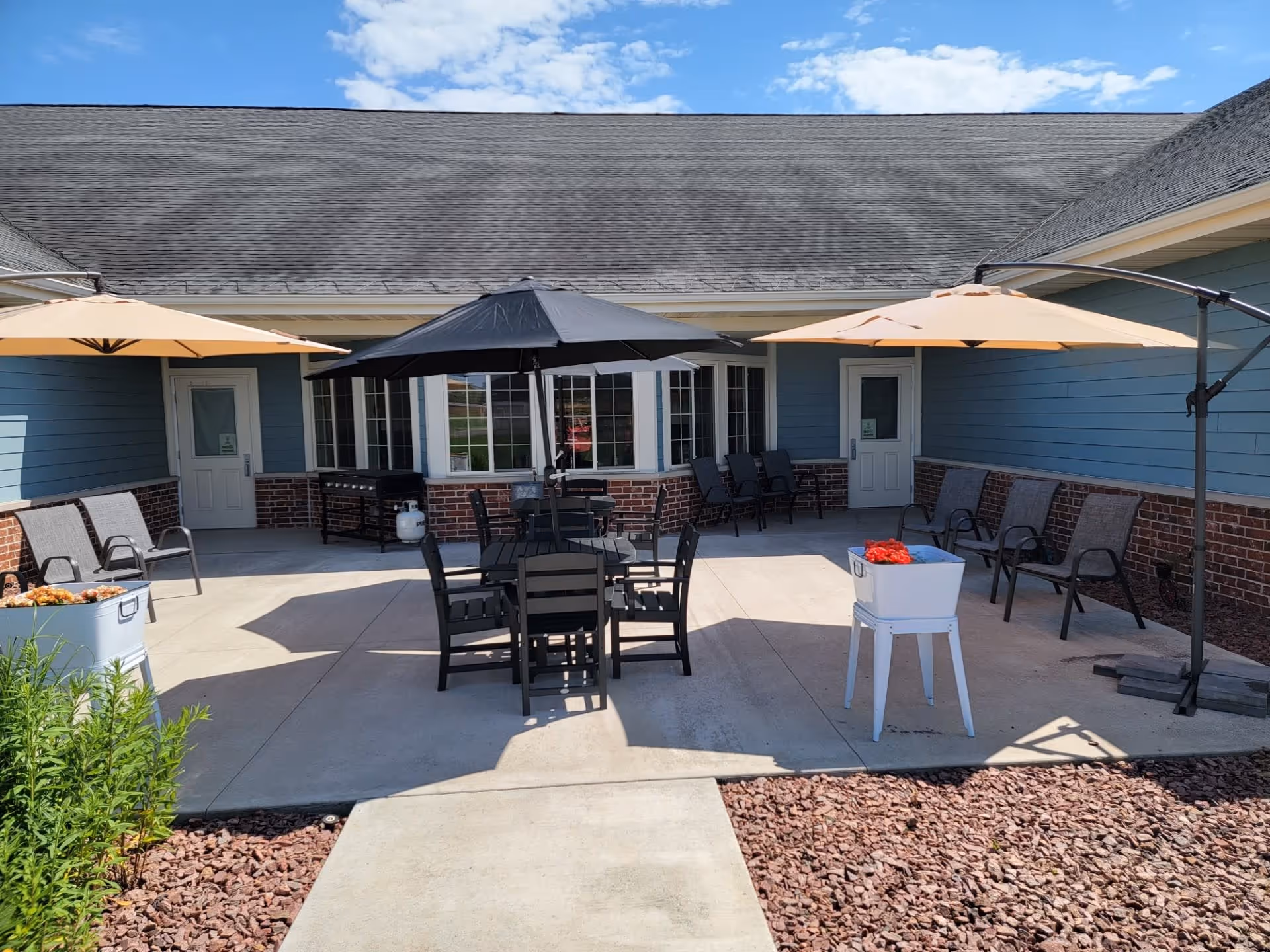 Outdoor patio area at The Waterford at Colby with several chairs arranged around tables shaded by large umbrellas. The patio is surrounded by a building with blue siding and brick accents, and there are two doors visible leading inside. The sky is clear with a few clouds.