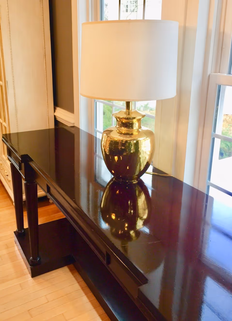 A shiny dark wooden console table with a reflective surface, holding a gold-colored lamp with a white lampshade, positioned in front of a window with white frames.